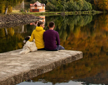 Couple sitting by the dock with their dog, looking at a cottage across the lake.