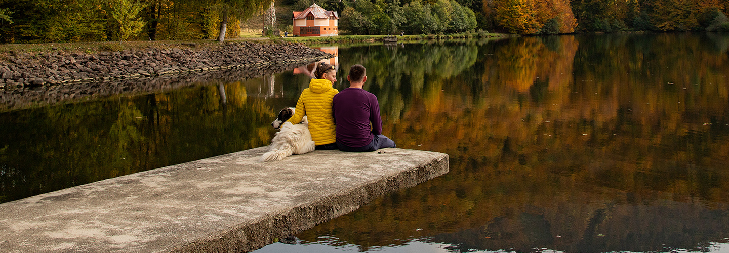 Couple sitting by the dock with their dog, looking at a cottage across the lake.